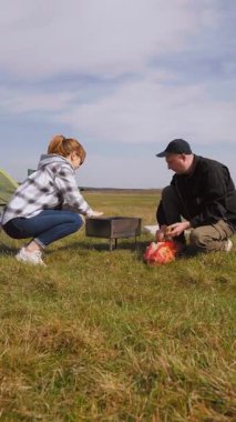 Genç adam çimenli bir alanda piknik yapmak için portatif barbekü ızgarası yapıyor. Kız arkadaşı da yakınlardaki katlanır sandalyede dinleniyor ve açık hava maceralarının keyfini çıkarıyor.