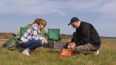 Kafkasyalı genç çift kamp alanlarında yemek pişirmek için barbekü ızgarası hazırlıyor. Yeşil bir çayırda çadır kurarak pikniğin tadını çıkartırken odunları baltaya koyuyorlar.