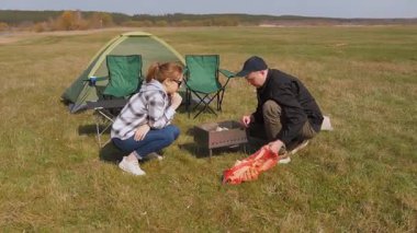 Kafkasyalı genç çift kamp alanlarında yemek pişirmek için barbekü ızgarası hazırlıyor. Yeşil bir çayırda çadır kurarak pikniğin tadını çıkartırken odunları baltaya koyuyorlar.