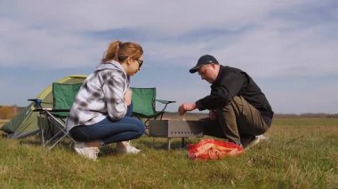 Kafkasyalı genç çift kamp alanlarında yemek pişirmek için barbekü ızgarası hazırlıyor. Yeşil bir çayırda çadır kurarak pikniğin tadını çıkartırken odunları baltaya koyuyorlar.