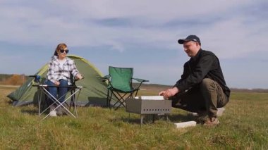 Genç adam yemek pişirmek için portatif barbekü ızgarası hazırlarken bir kadın güneşli bir günde açık hava pikniğinde çadırın yanındaki sandalyede dinleniyor.