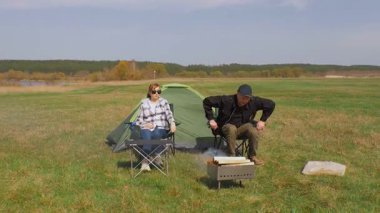 Olgun bir adam barbekü için ızgarada ateş yakarken bir kadın arka planda gölle birlikte yeşil bir çayırda piknik yaparken çadırın yanındaki katlanır sandalyede dinleniyor.