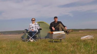 Genç çift çadırın yanındaki katlanır sandalyede oturur, güzel yeşil bir tarlada barbekü ızgarasında yemek pişirirken kırsal kesimde güneşli bir günde pikniğin tadını çıkarırlar.