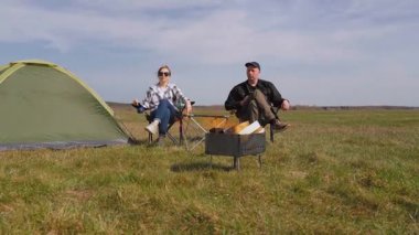 Genç çift çadırın yanındaki katlanır sandalyede oturur, güzel yeşil bir tarlada barbekü ızgarasında yemek pişirirken kırsal kesimde güneşli bir günde pikniğin tadını çıkarırlar.