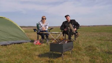 Genç çift çadırın yanındaki katlanır sandalyede oturur, güzel yeşil bir tarlada barbekü ızgarasında yemek pişirirken kırsal kesimde güneşli bir günde pikniğin tadını çıkarırlar.