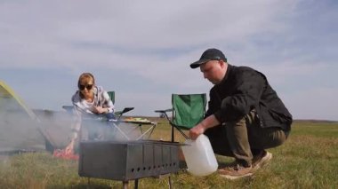 Genç bir adam, güneşli bir günde çayırda kamp yaparken arka planda bir masada oturan bir kadın teneke kutudan suyla barbeküyü söndürüyor.