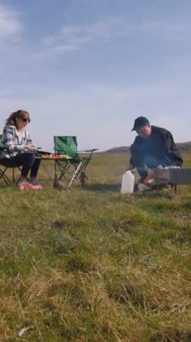 Genç bir adam barbeküde şiş kebap pişirirken kız arkadaşı da çadırlarının yanındaki çimenlik alanda kamp koltuğunda rahatlıyor ve birlikte pikniğin tadını çıkarıyor.