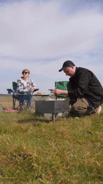 Genç bir adam barbeküde şiş kebap pişirirken kız arkadaşı da çadırlarının yanındaki çimenlik alanda kamp koltuğunda rahatlıyor ve birlikte pikniğin tadını çıkarıyor.