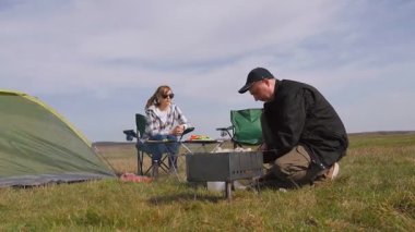 Genç çift doğada pikniğin tadını çıkarırken, erkek portatif bir ızgarada lezzetli bir barbekü hazırlarken partneri çimenli bir tarlada çadırlarının yanında dinleniyor.