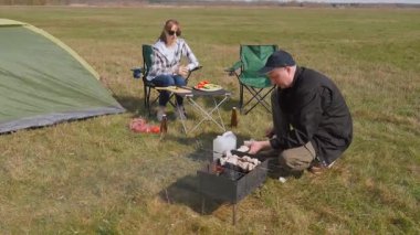 Genç çift doğada pikniğin tadını çıkarırken, erkek portatif bir ızgarada lezzetli bir barbekü hazırlarken partneri çimenli bir tarlada çadırlarının yanında dinleniyor.
