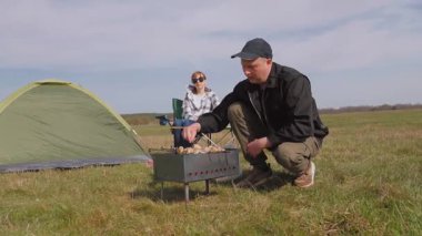 Genç adam ızgarada lezzetli şiş kebap pişirirken kız arkadaşı da çadırlarının yanındaki çimenlik alanda kamp koltuğunda dinleniyor ve birlikte pikniğin tadını çıkarıyor.