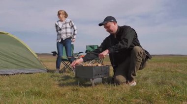 Genç adam ızgarada lezzetli şiş kebap pişirirken kız arkadaşı da çadırlarının yanındaki çimenlik alanda kamp koltuğunda dinleniyor ve birlikte pikniğin tadını çıkarıyor.
