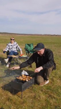 Beyzbol şapkalı genç bir adam güneşli bir günde arkadaşlarıyla açık hava pikniğinde şiş kebap pişirip şiş pişirip şiş kebap pişiriyor ve sıcak kömür üzerinde terbiye edilmiş et pişiriyor.