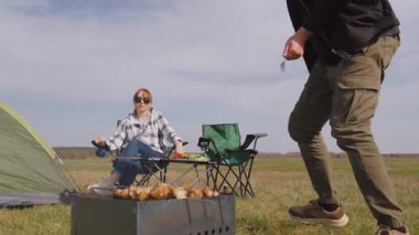 Genç çift doğada harika bir piknik yapıyor, barbeküde leziz et şişleri pişiriyor güneşli bir günde çadırlarının yanında kamp sandalyelerinde dinleniyorlar.