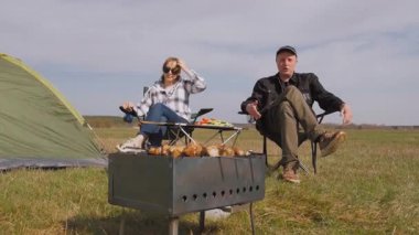 Genç adam ızgarada lezzetli şiş kebap pişirirken kız arkadaşı da çadırlarının yanındaki çimenlik alanda kamp koltuğunda dinleniyor ve birlikte pikniğin tadını çıkarıyor.