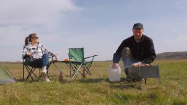 Genç adam ızgarada lezzetli şiş kebap pişirirken kız arkadaşı da çadırlarının yanındaki çimenlik alanda kamp koltuğunda dinleniyor ve birlikte pikniğin tadını çıkarıyor.