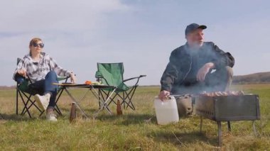 Genç adam ızgarada lezzetli şiş kebap pişirirken kız arkadaşı da çadırlarının yanındaki çimenlik alanda kamp koltuğunda dinleniyor ve birlikte pikniğin tadını çıkarıyor.