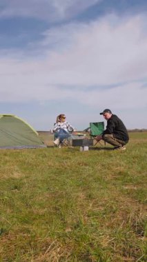 Mutlu çift kömür ızgarasında leziz ızgara yaparken manzaralı yeşil alanda piknik yaparken güneşli bir günde birlikte açık hava maceralarının keyfini çıkarırken ve kamp çadırlarının yanında dikey video görüntüleriyle birlikte.