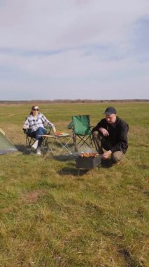Genç bir adam barbeküde şiş kebap pişirirken kız arkadaşı da çadırlarının yanındaki çimenlik alanda kamp koltuğunda rahatlıyor ve birlikte pikniğin tadını çıkarıyor.