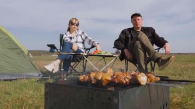 Genç çift konuşurken ve eğlenirken çadırın yanındaki sandalyeleri katlarken ve bir tarladaki piknik sırasında şiş kebap üzerinde şiş kebap eti pişirirken.