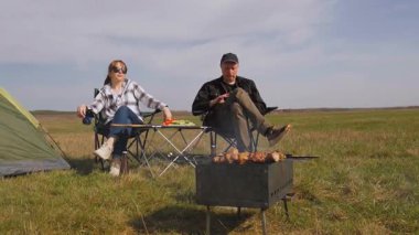 Genç adam ızgarada lezzetli şiş kebap pişirirken kız arkadaşı da çadırlarının yanındaki çimenlik alanda kamp koltuğunda dinleniyor ve birlikte pikniğin tadını çıkarıyor.