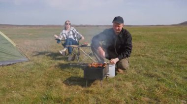 Olgun bir adam portatif bir barbeküde et şişi pişirirken bir kadın güneşli bir günde huzurlu bir kamp pikniğinde çadırın yanındaki katlanır sandalyede dinleniyor.