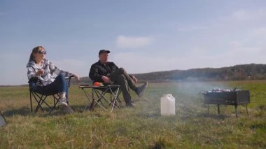 Genç çift çadırın yanındaki katlanır sandalyede oturur, güzel yeşil bir tarlada barbekü ızgarasında yemek pişirirken kırsal kesimde güneşli bir günde pikniğin tadını çıkarırlar.