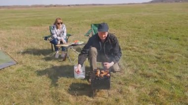 Mutlu çift kömür ızgarasında leziz ızgara yaparken manzaralı yeşil alanda piknik yaparken güneşli bir günde kamp çadırının yanında birlikte maceralarının tadını çıkarıyorlar.