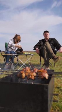 Beyzbol şapkalı genç bir adam güneşli bir günde arkadaşlarıyla açık hava pikniğinde şiş kebap pişirip şiş pişirip şiş kebap pişiriyor ve sıcak kömür üzerinde terbiye edilmiş et pişiriyor.