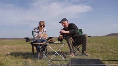 Açık havada güneşli bir günün tadını çıkaran genç bir çift, çadırın yanındaki sandalyelere oturmuş, güzel mavi gökyüzünün altındaki yeşil bir çayırda piknikte ızgara yemek yiyor ve sohbet ediyorlar.