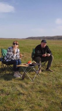 Açık havada güneşli bir günün tadını çıkaran genç bir çift, çadırın yanındaki sandalyelerde oturmuş, güzel mavi gökyüzü dikey videosunun altında yeşil bir çayırda piknikte ızgara yemek yiyor ve sohbet ediyorlar.