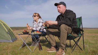 Açık havada güneşli bir günün tadını çıkaran genç bir çift, çadırın yanındaki sandalyelere oturmuş, güzel mavi gökyüzünün altındaki yeşil bir çayırda piknikte ızgara yemek yiyor ve sohbet ediyorlar.