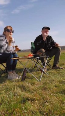 Açık havada güneşli bir günün tadını çıkaran genç bir çift, çadırın yanındaki sandalyelerde oturmuş, güzel mavi gökyüzü dikey videosunun altında yeşil bir çayırda piknikte ızgara yemek yiyor ve sohbet ediyorlar.