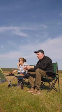 Açık havada güneşli bir günün tadını çıkaran genç bir çift, çadırın yanındaki sandalyelerde oturmuş, güzel mavi gökyüzü dikey videosunun altında yeşil bir çayırda piknikte ızgara yemek yiyor ve sohbet ediyorlar.