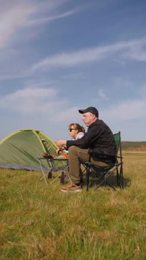 Açık havada güneşli bir günün tadını çıkaran genç bir çift, çadırın yanındaki sandalyelerde oturmuş, güzel mavi gökyüzü dikey videosunun altında yeşil bir çayırda piknikte ızgara yemek yiyor ve sohbet ediyorlar.