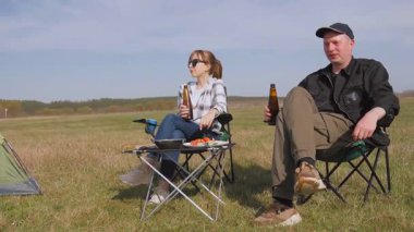 Genç çift kamp sandalyelerinde oturuyor, bira içiyor, konuşuyor ve güneşli bir günde güneşli bir alanda barbekü pikniğinin tadını çıkarıyor.