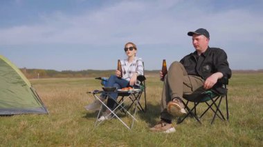 Genç çift kamp sandalyelerinde oturuyor, bira içiyor, konuşuyor ve güneşli bir günde güneşli bir alanda barbekü pikniğinin tadını çıkarıyor.