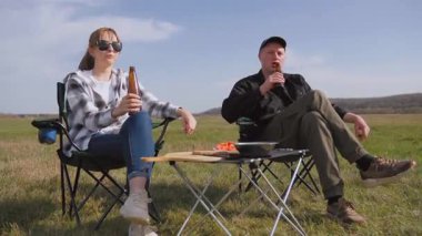 Genç çift kamp sandalyelerinde oturuyor, bira içiyor, konuşuyor ve güneşli bir günde güneşli bir alanda barbekü pikniğinin tadını çıkarıyor.