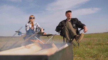 Genç adam ve kadın katlanır sandalyelerde oturur, engin yeşil bir tarlada barbeküyle pikniğin tadını çıkarırken, kadın bira içer ve güneşli bir günde birlikte dinlenir.