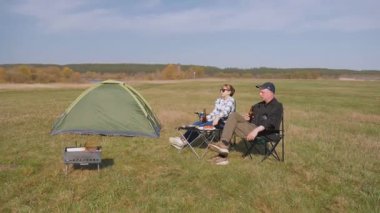 Genç çift, güneşli bir günde güneşli bir çayırda güneşli bir piknik ve barbekü yaparak, kamp alanında, katlanan sandalyelerde rahatlıyor ve bira içiyor.