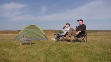 Genç çift, güneşli bir günde güneşli bir çayırda güneşli bir piknik ve barbekü yaparak, kamp alanında, katlanan sandalyelerde rahatlıyor ve bira içiyor.