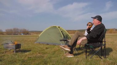 Neşeli bir çift piknik yaparken çadırlarının yanında bira, katlanabilir sandalyelerde dinlenirken ve güneşli bir bahar gününde yeşil bir çayırda sohbet ederken eğleniyor.