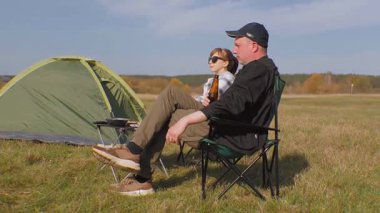 Genç çift, güneşli bir günde güneşli bir çayırda güneşli bir piknik ve barbekü yaparak, kamp alanında, katlanan sandalyelerde rahatlıyor ve bira içiyor.