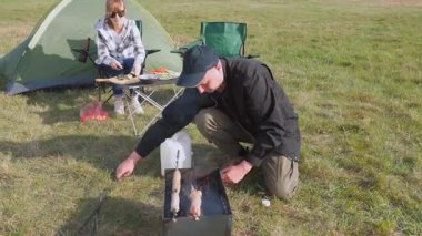Genç adam portatif kömür ızgarasında leziz et kebabı hazırlarken kız arkadaşı güneşli bir günde kamp alanında salata için sebze kesiyor.