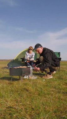Mutlu yetişkin çift güneşli bir günde pikniğin keyfini çıkarırken, erkek barbeküde sosis pişirirken kadın güzel yeşil bir çayırda kamp masasında oturuyor.