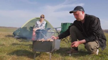 Genç adam portatif kömür ızgarasında leziz et kebabı hazırlarken kız arkadaşı güneşli bir günde kamp alanında salata için sebze kesiyor.