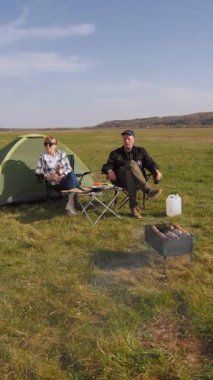 Mutlu bir adam ve kadın çadırlarının yanındaki kamp sandalyelerinde dinleniyor. Güneşli bir günde çimenli bir tarlada dikey bir videoda şiş kebabıyla barbekü pikniğinin keyfini çıkarıyorlar.