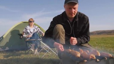 Güneşli bir günde, doğada pikniğin tadını çıkaran genç bir çift, dumanlı bir barbekü üzerinde et ızgarası yapan bir adam, bir kadın, yeşil bir çadırın yanında kamp koltuğunda dinlenirken,