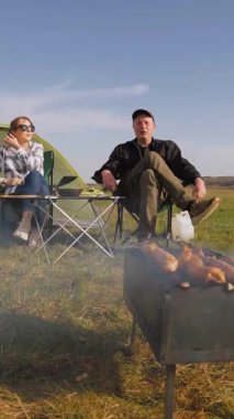 Mutlu bir adam ve kadın çadırlarının yanındaki kamp sandalyelerinde dinleniyor. Güneşli bir günde çimenli bir tarlada dikey bir videoda şiş kebabıyla barbekü pikniğinin keyfini çıkarıyorlar.