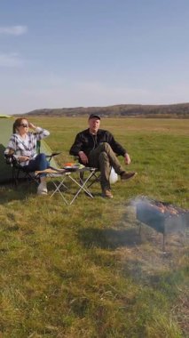 Mutlu bir adam ve kadın çadırlarının yanındaki kamp sandalyelerinde dinleniyor. Güneşli bir günde çimenli bir tarlada dikey bir videoda şiş kebabıyla barbekü pikniğinin keyfini çıkarıyorlar.